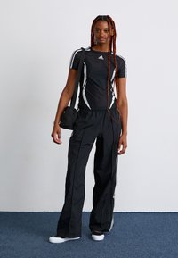 Young woman with long braids wearing black Adidas athletic shirt and pants, carrying black shoulder bag, standing on blue carpet.