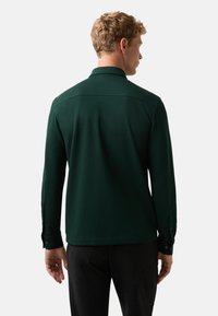 Mann mit kurzen lockigen blonden Haaren, der ein dunkelgrünes Langarmshirt und dunkle Hosen trägt, steht mit dem Rücken zu einer einfarbigen Hintergrund.