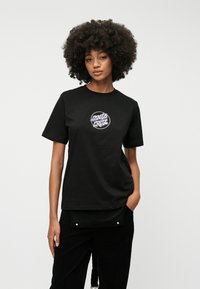 Μαύρη βαμβακερή μπλούζα με στρογγυλό λευκό και μωβ λογότυπο εκτύπωσης. Διαθέτει κοντά μανίκια και χαλαρή εφαρμογή. Φοράει με μαύρες βελούδινες παντελόνες.