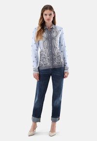 Camicia azzurra chiara button-down con motivo paisley grigio, abbinata a jeans denim scuri arrotolati ai polsini e scarpe con tacco beige.