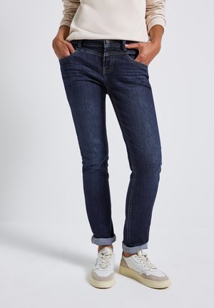 Jean en denim bleu foncé avec une coupe slim, présentant un design classique à cinq poches et des ourlets retroussés, associés à des baskets blanches et beiges.