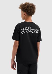 Jongen met krullend haar, gekleed in een zwart t-shirt met witte 3D-stijl tekst "BILLINI" op de achterkant, staand tegen een effen lichte achtergrond.