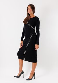 Abito midi nero a coste con maniche lunghe, caratterizzato da una fila diagonale di accessori in filo di argento lungo un lato. Texture liscia, forma aderente.