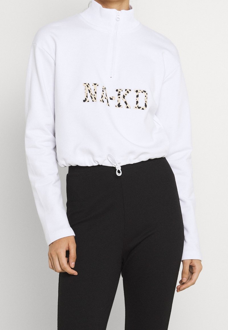 Sudadera blanca con cremallera y cordón en el dobladillo, con un logotipo de leopardo "NA-KD" en el frente. Combinada con pantalones negros ajustados.
