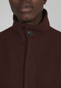 Burgundi gyapjúszövet kabát magas gallérral és egy fekete gombbal záródik, puha textúrájú és letisztult, áramvonalas dizájnnal rendelkezik.