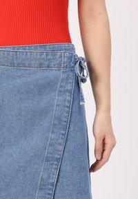 Denim omslagskjørt med lyseblått stoff, med en knytespenne på siden. Stoffet har en glatt tekstur med minimal detaljering.