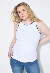 Witte tanktop met zwarte decoratieve afwerking rond de halslijn en armgaten, gecombineerd met blauwe spijkerbroek. Geribbeld textuur. Effen achtergrond.