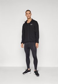 Mustan vetoketjullinen huppari, jossa on istuva malli, päällä mustat legginssit. Rinnassa on logo ja asusteena mustat urheilukengät.