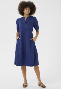 Tamsiai mėlyna suknelė su trumpomis pūstomis rankovėmis, sagomis užsegama iškirpte, šoninėmis kišenėmis ir plazdančiu kraštu. Lygus audinio paviršius.