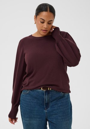 Mulher com cabelo escuro apanhado num coque, vestindo um casaco roxo de manga comprida e jeans azuis, tocando o pescoço e olhando para baixo.