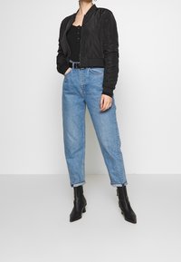 Černá bomber bunda s žebrovanými manžetami přes černý tílko, zkombinovaná s džínami s vysokým pasem ve světle modré barvě a černými kotníkovými botami.