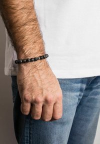 Brăță din mărgele negre pe un încheietură, având mărgele sferice, lucioase și mate, piele texturată și o cămașă albă simplă.