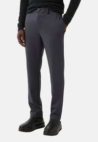 Grijze, rekbare broeken met een gladde textuur, aansluitende pasvorm, voorzakken en een enkele knoopsluiting. Gecombineerd met zwarte schoenen.