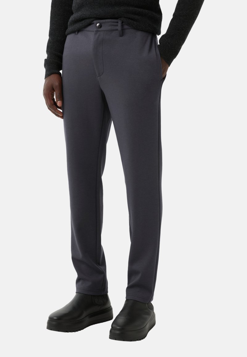 Grijze, rekbare broeken met een gladde textuur, aansluitende pasvorm, voorzakken en een enkele knoopsluiting. Gecombineerd met zwarte schoenen.