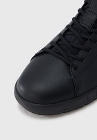 Μαύρο δερμάτινο αθλητικό παπούτσι με υφή στο μέρος της μύτης, μαύρα κορδόνια και παχύ υφασμάτινο πέλμα σε ουδέτερο φόντο.