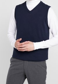 Marineblaues ärmelloses Strickweste mit V-Ausschnitt, getragen über einem weißen Langarmshirt, gezeigt an einem Modell mit verschränkten Händen.