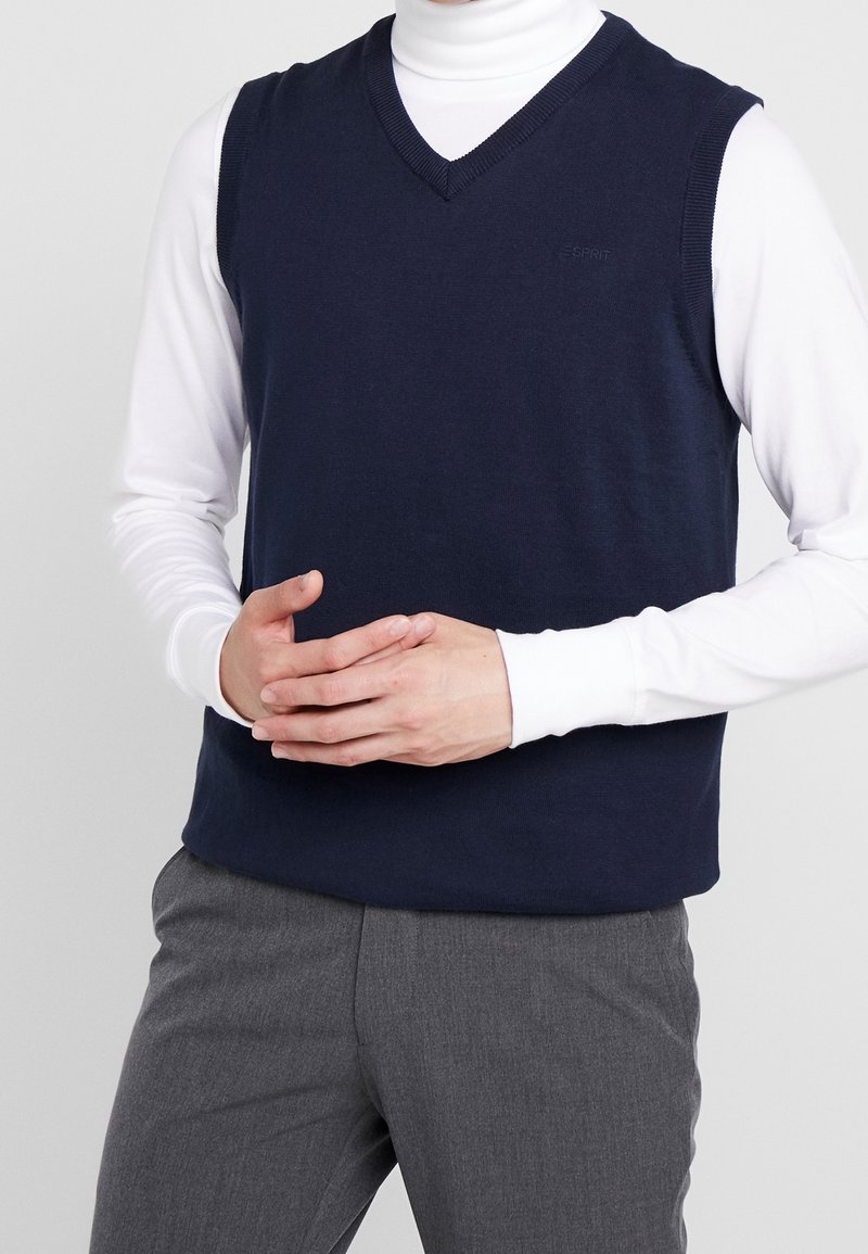 Marineblaues ärmelloses Strickweste mit V-Ausschnitt, getragen über einem weißen Langarmshirt, gezeigt an einem Modell mit verschränkten Händen.