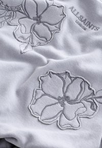 Tessuto grigio con un intricato ricamo floreale in grigio chiaro. Il testo dice "ALLSAINTS LONDON." Texture morbida con una finitura liscia.