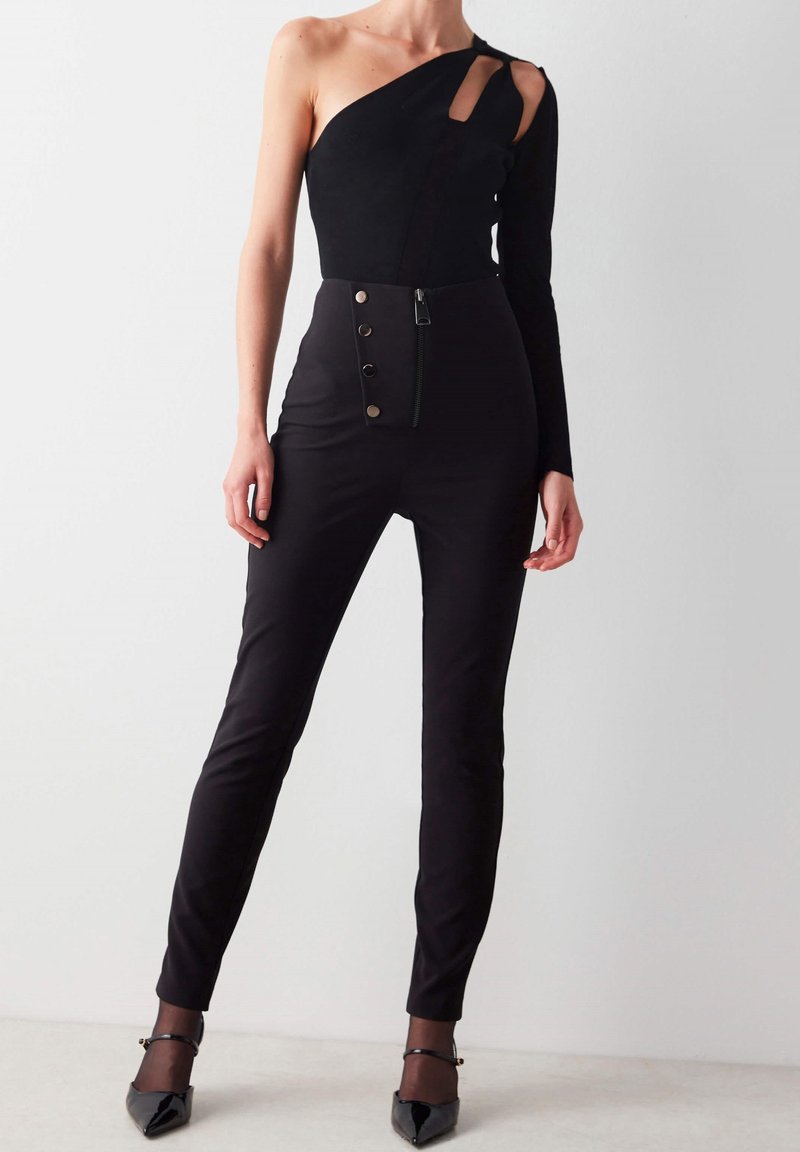 Femme portant une combinaison noire asymétrique à une manche avec taille haute, boutons visibles et escarpins noirs pointus, debout devant un mur uni.