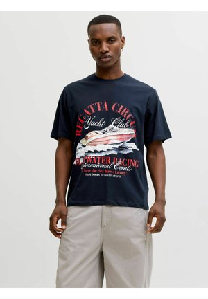 Fiatal férfi tengerészkék mintás pólóban, rajta piros-fehér vitorlásverseny motívum, bézs rövidnadrágban, egyszínű világos háttér előtt.