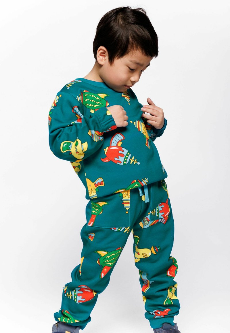 Niño pequeño con pantalones de chándal de color teal con estampados coloridos y una camiseta de manga larga, mirando hacia abajo a su ropa con una expresión pensativa.