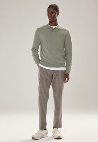 Lichtgroene trui met col, zacht materiaal; gecombineerd met bruine gestreepte broek en witte sneakers. Simpele, aansluitende stijl.