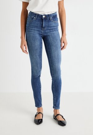 Kobieta ubrana w niebieskie rurki z wysokim stanem, biały top i czarne, spiczaste baleriny Mary Jane, stojąca na jednolitym białym tle.