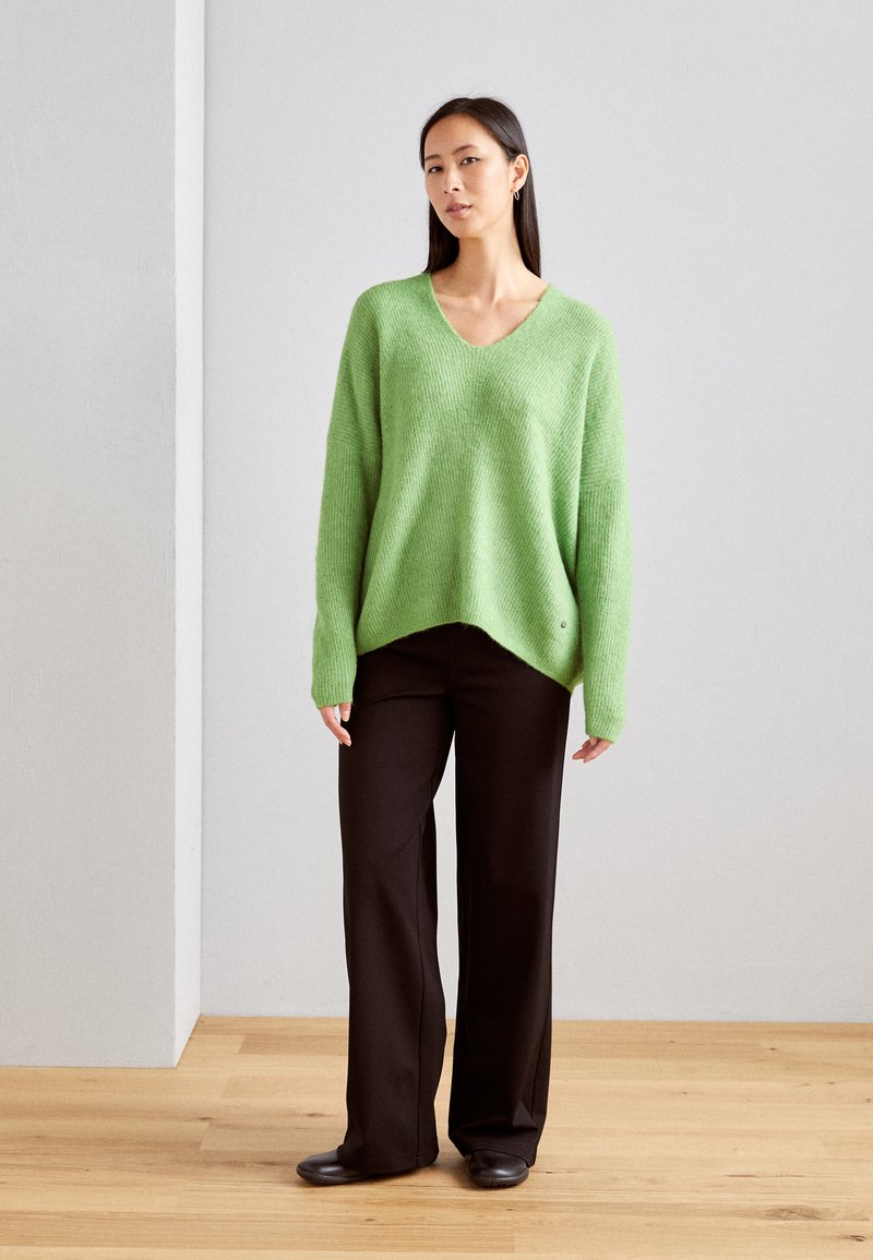 Maglione verde a V realizzato in materiale soffice, indossato sopra pantaloni neri a gamba larga. Il maglione presenta spalle calate e un orlo leggermente asimmetrico.
