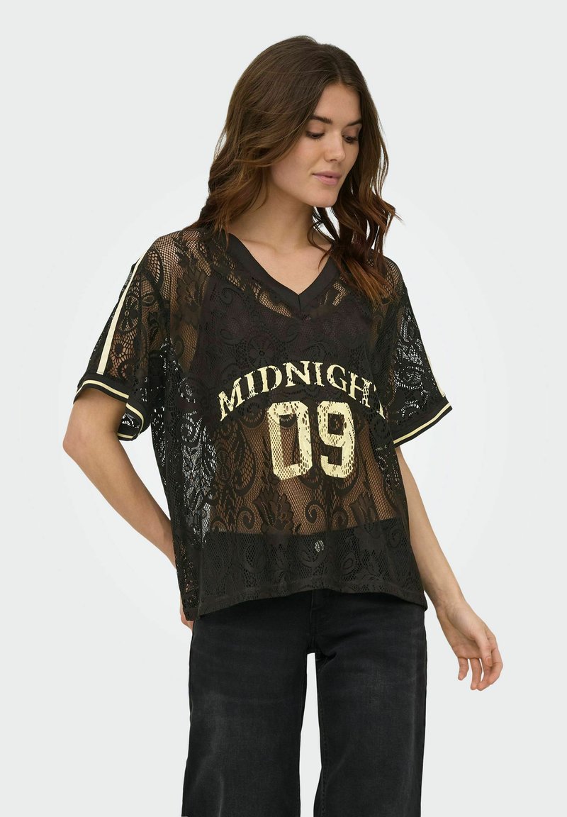 Jeune femme aux longs cheveux bruns portant un haut noir en dentelle à manches courtes avec l’inscription "MIDNIGHT 09" et un pantalon noir, debout, une main dans le dos.