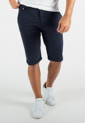 Shorts bleu marine en tissu, avec des ourlets roulés, des poches latérales et une fermeture discrète. Associés à des baskets blanches.