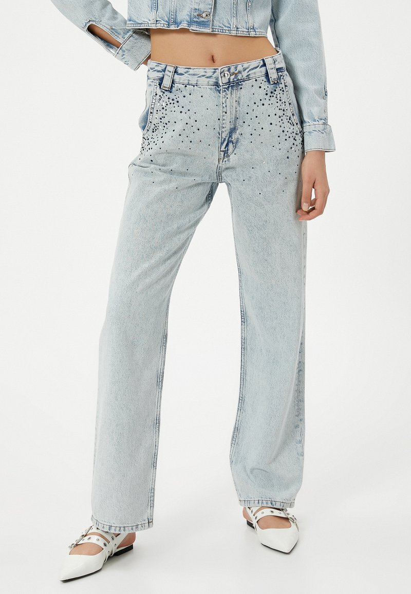 Vaqueros de pierna ancha de denim azul claro con una cintura alta, decorados con tachuelas plateadas en la parte delantera. Textura suave con un acabado desvanecido.