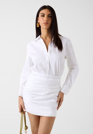Mujer con cabello largo y oscuro, lleva una camisa blanca abotonada y una minifalda blanca fruncida, sosteniendo un bolso verde con asa de cuerda sobre fondo liso.