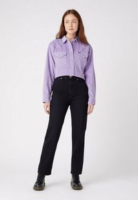 Fioletowa sztruksowa krótka koszula z zapięciem na guziki i dwiema kieszeniami na piersi, w połączeniu z czarnymi jeansami o wysokim stanie i prostych nogawkach oraz czarnymi botkami.