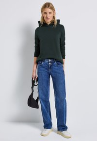 Hoodie vert foncé avec un capuchon à cordon, associé à un jean en denim bleu. L'ensemble présente une coupe décontractée et des baskets blanches.