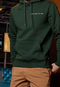Green hoodie made of soft fabric with a front pocket, features white text "NEW YORK, EST. 1985" on the chest, adjustable drawstring hood.