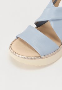 Sandale en cuir bleu clair avec des brides croisées, une semelle intérieure lisse et une semelle beige épaisse avec une garniture métallique tressée.