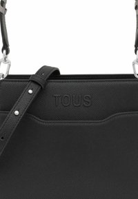 Czarna skórzana torba na ramię o teksturowanym wykończeniu, wytłoczone logo "TOUS", regulowany pasek, srebrne akcenty metalowe oraz minimalistyczny design.