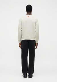 Ανοιχτό γκρι φούτερ με στρογγυλή λαιμόκοψη, μανίκια με μανσέτες και μικρό πορτοκαλί λογότυπο στο πίσω μέρος, συνδυασμένο με φαρδιά μαύρα τζιν και μαύρα παπούτσια.