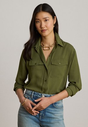 Femme aux longs cheveux foncés portant une chemise kaki à boutons, un jean bleu, un collier en chaîne dorée, un bracelet et de petites boucles d'oreilles, se tenant détendue.