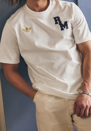 Personne portant un T-shirt blanc avec logo Adidas doré et lettres RM noires, pantalon beige, bracelet argenté et cheveux bouclés visibles.