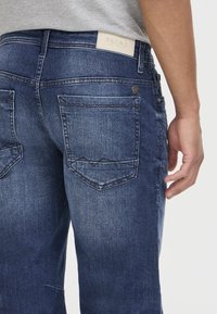 Jeans de mezclilla azul oscuro con un acabado texturizado, que cuentan con bolsillos traseros y una etiqueta de marca de cuero en la cintura.