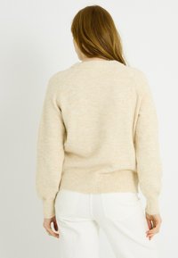 Suéter beige de punto con un corte relajado, mangas raglán y dobladillo acanalado. Textura suave y variaciones sutiles de color. Se lleva con pantalones blancos.