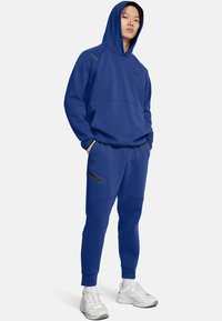Joven con una sudadera con capucha azul y pantalones jogger a juego, con zapatillas blancas, de pie con las manos en los bolsillos.
