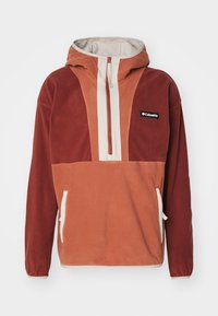 Pullover Columbia in arancione ruggine e rosso mattone, con una zip frontale a metà, cappuccio, tasche laterali e un tessuto morbido e testurizzato.