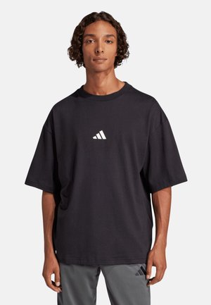 Giovane uomo con capelli ricci che indossa una maglietta nera larga Adidas e pantaloni grigi con un logo Adidas sulla coscia.