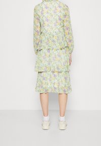 Žena stojící zády, oblečená v vrstevnatých pastelových květinových šatech s dlouhými rukávy, v bílých ponožkách a bílých teniskách na jednobarevném pozadí.