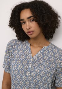 Chemise à motifs bleu et crème avec des manches courtes, dotée d'un col en v et de détails en boutons. Fabriquée dans un tissu léger avec un design floral.