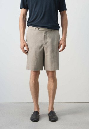 Mann trägt beige knielange Shorts, schwarze Schlupfschuhe und ein dunkelblaues T-Shirt mit kurzen Ärmeln, steht vor einfachem Hintergrund.