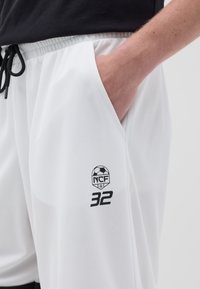 Shorts deportivos blancos hechos de tela de malla, con bolsillos laterales, un cordón negro y un logo impreso con el texto "NCF 32" en el muslo.