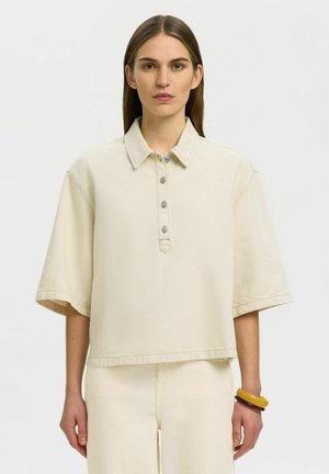 Kobieta o prostych brązowych włosach, ubrana w za dużą, ecru koszulę z krótkim rękawem i kołnierzykiem oraz pasujące spodnie, z bransoletkami na nadgarstku.